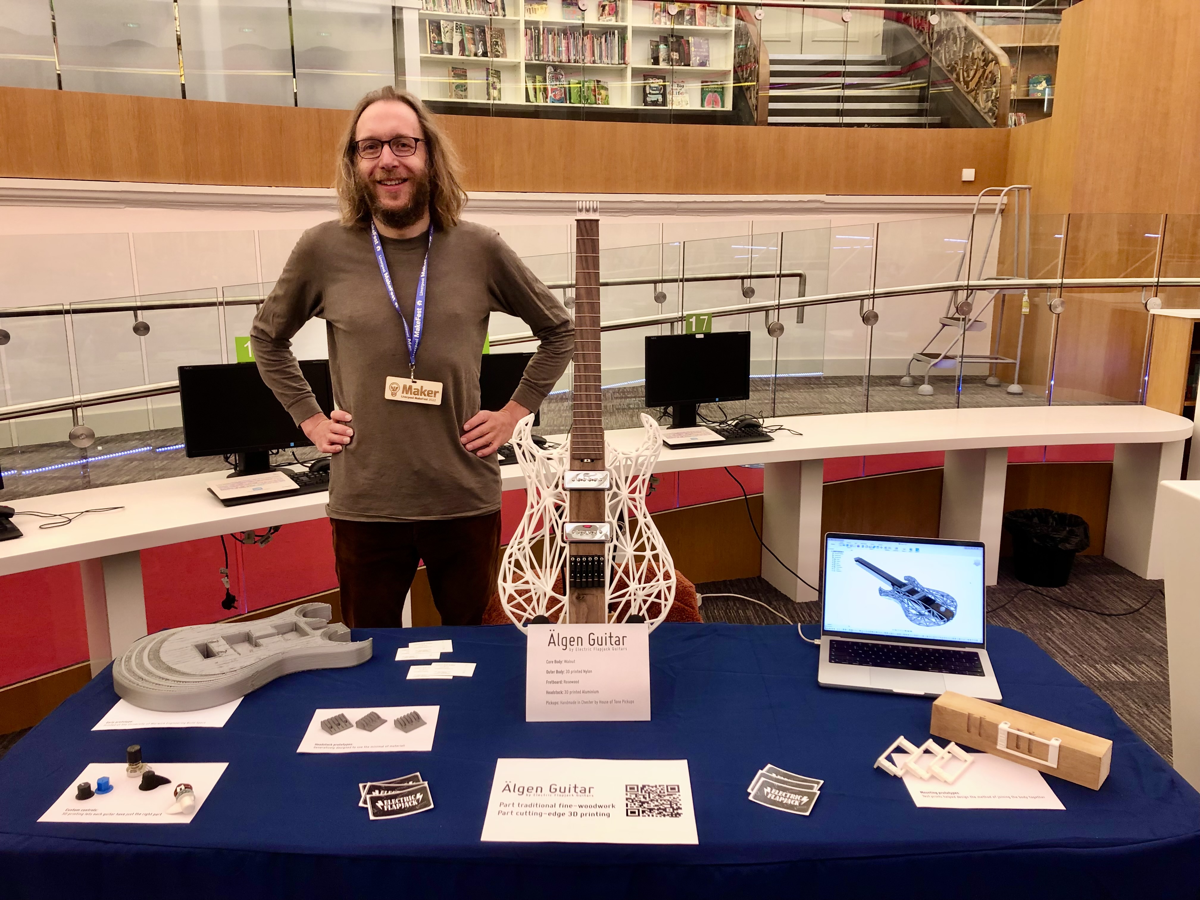 A photo of me stood behind a table at a show. On the table is a close to finished Älgen guitar (but not yet finished as there's no strings on it). On the table around the guitar are a selection of prototype parts and a laptop showing the CAD model.