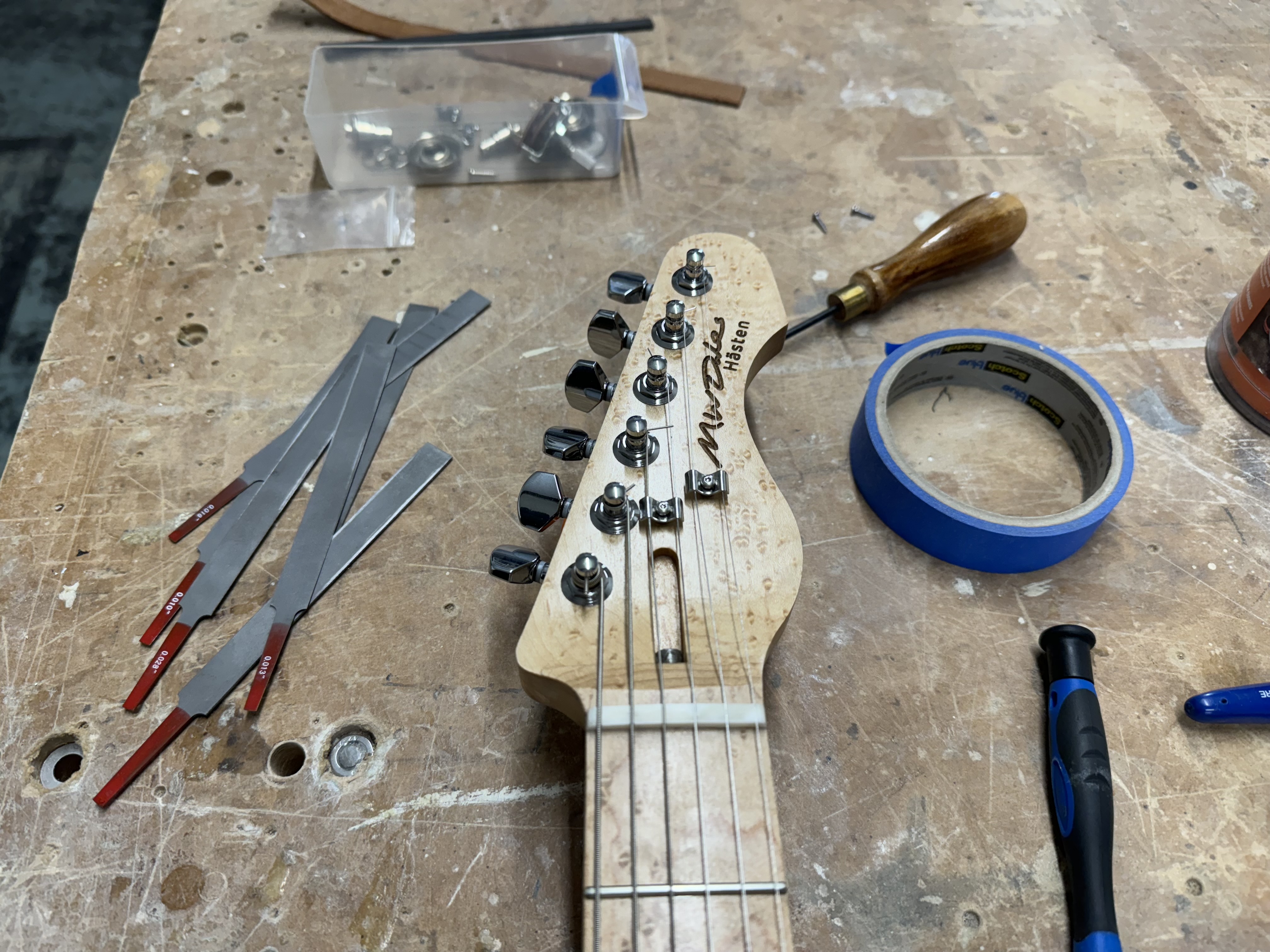 A close up photo of the headstock of the guitar, showing the brids-eye maple in detail. You can see the tuners, the nut and the two strings trees (along with the strings). Laser etched into the headstock is 'M W Dales' and 'Hästen'. Next to the headstock on the workbench are a set of tiny files for cutting slots into the nut.