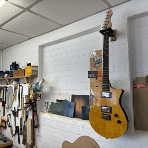 A photo of a completed guitar hung on the wall in the workshop, showing a body with birds-eye maple cap stained bright yellow, and black sides, with a maple neck and a rosewood fretboard. It has a chrome covered P90 in the neck position and a chrome covered humbucker in the bridge position.