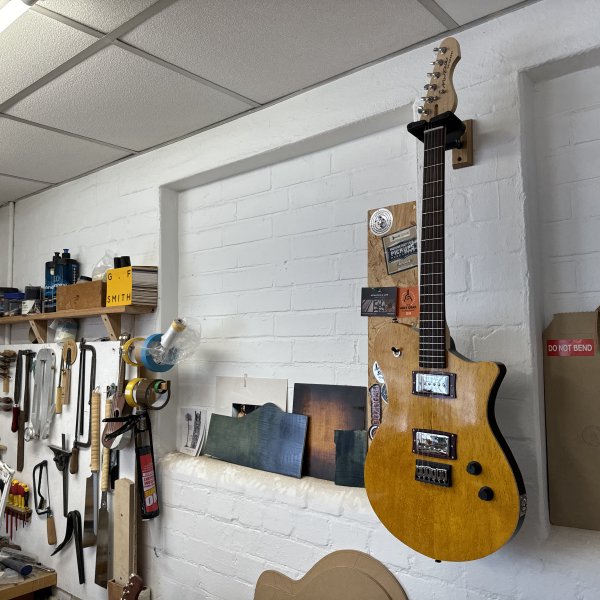 A photo of a completed guitar hung on the wall in the workshop, showing a body with birds-eye maple cap stained bright yellow, and black sides, with a maple neck and a rosewood fretboard. It has a chrome covered P90 in the neck position and a chrome covered humbucker in the bridge position.